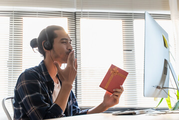 Happy young Asian man showing gift box while having video call on computer with his girlfriend or family at home. Distance relations, New normal celebrations during Coronavirus pandemic.