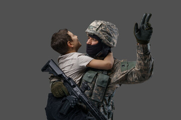 Portrait of soldier patriot dressed in camouflage uniform hugging kid against grey background.