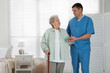 © New Africa - Caregiver assisting senior woman with walking cane indoors. Home health care service