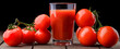 A glass cup with tomato juice, next to it lies a branch of tomatoes. On a wooden table there is a glass of tomato juice and tomatoes lie on a black background.