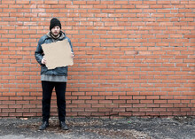 Homeless Holds Piece Of Cardboard. Refugee Is At Brick Wall Looking For Job. Male Tramp In Dirty Clothes. Below Poverty Line. Bearded Fugitive Beggar. Drunk Immigrant. Copy Space For Text