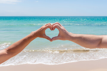 Hands forming a heart in front of a beach of golden sand and turquoise water. Love in paradise