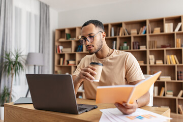 Poster - Middle eastern male student learning remotely, reading book, browsing internet on laptop and drinking coffee at home