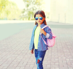 Wall Mural - Stylish little girl child wearing sunglasses, shirt with pink backpack on city street background