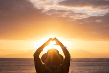 Rear View Of Caucasian Senior Woman At Golden Sunset Standing At The Beach Gesturing Heart Shape With Hands Looking At Horizon Over Water. Relaxed Elderly White Haired Lady Enjoying Sea Vacation