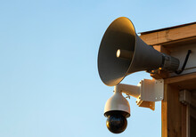 Camera, Loudspeaker On A Wall Of A Building Against Blue Sky. CCTV Security Video Camera With Loudspeakers And Megaphone Isolated On Blue Background. Webcam Overlook A Public Area In Sea. Copy Space