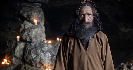 Wall Mural - medieval monk, with a staff in a hooded place, stands in a dark cave with glowing candles in the bac