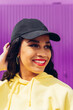 © Dani D.G - Portrait of the face of a smiling young African woman, in a yellow cap and sweatshirt on a purple background.Young girl with reggaeton and hip hop style urban clothes.