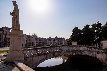 Woman in a dress and hat sitting on bridge of Prato della Valle, square in the city of Padua, Veneto, Italy, Europe. Isola Memmia surrounded by canal bordered by two rings of statues. Reflection water