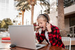 © Elena K - A blond schoolboy in a plaid red shirt sits at a table with headphones and a laptop.  A smiling boy rejoices in victory.  Distance learning. Homeschooling.  Video communication.