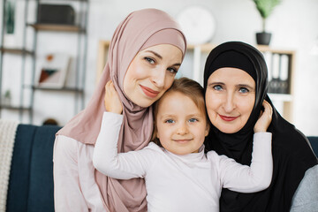 Head shot portrait happy three generations of islamic women in hijab posing for family picture, smiling mature islam grandmother and mother with adorable little girl looking at camera.