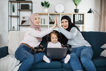 Wall Mural - Three generations of muslim women look at camera while playing video games at home, sitting on sofa with laptop and using joysticks. Islamic mother and grandmother make a high five to win the game.