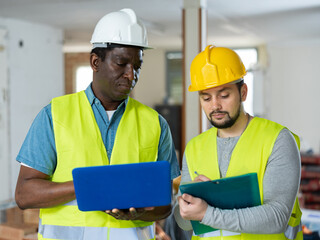 Two skilled maintenance workers wearing protective helmets and vests talking work progress and gesturing at indoor building site