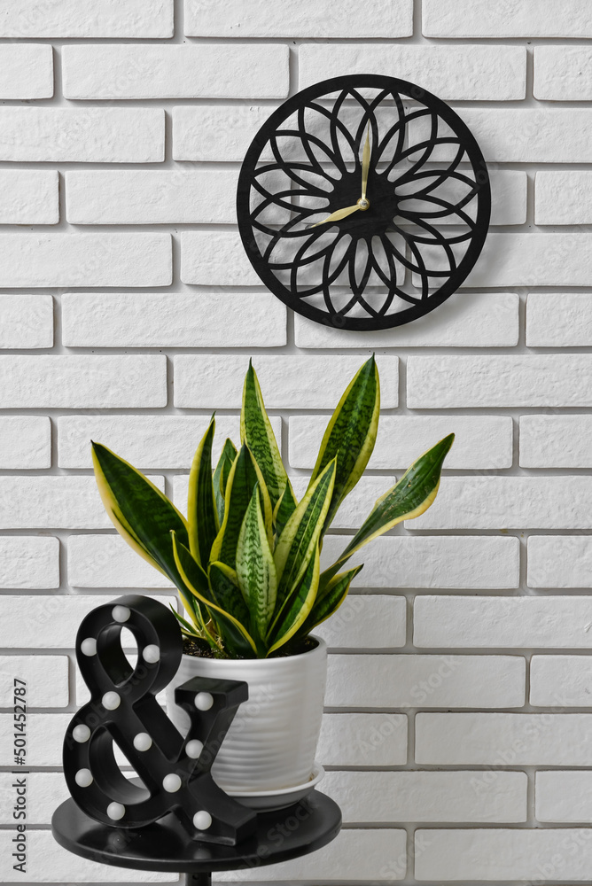 Table with houseplant and modern clock hanging on light brick wall in room