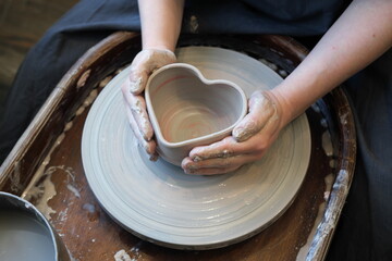 Women hands on a potter wheel embrace a heart-shaped pot.