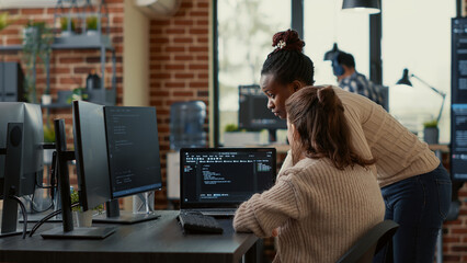 Two system engineers analyzing source code on laptop looking for errors on screen while sitting at desk. Team of app developers working on group project for ai online cloud computing project.