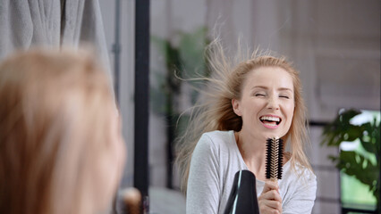 Wall Mural - Caucasian young woman in Ukraine blowing her hair with a hair dryer and singing with a brush