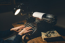 Typing Hands Of A Writer Looking For Inspiration To Start A New Novel On His Vintage Typewriter In A Retro Style Studio: A Wooden Desk Lit By A Lamp, Old Books And A Pair Of Glasses.