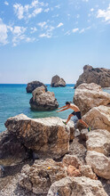 A Young Man Jumping Between Sharp Rocks In Aphrodite's Birthplace, Petra Tou Romiou, Cyprus.  Sea Is Calm, In Turquoise Shade. Other Smaller Boulders Emerging From The Sea. Freedom And Relaxation