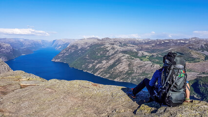 a man wearing blue shirt, full cap and a huge hiking backpack sits at the edge of a steep cliff of p