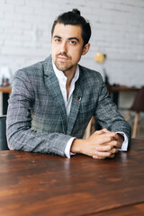 Wall Mural - Vertical portrait of confident stylish business man in fashion suit sitting at desk in modern office room, looking at camera. Front view of serious bearded businessman posing at workplace.