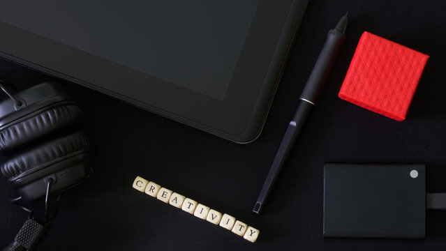Stylus and graphics tablet next to external ssd drive, red box, headphones and the inscription creativity. Black background. Сoncept of modern computer creativity, design and digital devices