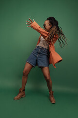 Wall Mural - Emotional african girl in shorts and shirt dancing, having fun isolated on dark green background. Concept of beauty, art, fashion, youth and emotions