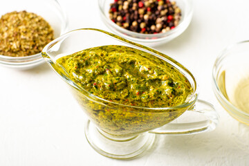 Chimichurri sauce in a gravy bowl on a white background. Various spices lie nearby. Argentinean vegetarian sauce made from olive oil, oregano, parsley.