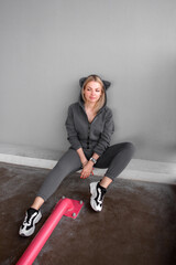 Wall Mural - teen leisure. blonde girl in gray oversize overalls with hood with kitty ears sits casual at parking near gray wall background and looks apart with cute smile. sport lifestyle concept, free space