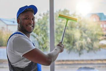 Cleaning plastic windows against background of courtyard of house with green trees and sun by smiling black worker. Handsome young worker maintains order in office, gladly performs household chores