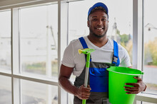 Employee Cleaning Service In Overalls And Blue Baseball Cap Happily Poses Against Background Of Clean Window. Perfectly Washed Window Sparkles In Sun, Dark-skinned Cleaner Is Happy With Job Done