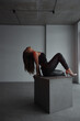 © NASTYA PALEHINA - young brunette woman in black top and leather pants is sitting fashion in profile on the gray cube and looking up on the white wall background in loft interior. fashion concept, free space