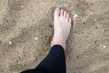 Man Foot On The Beach In Rocky Sand Wearing A Black Wetsuit Before Surfing. White Male Foot In Beach Sand Before Water Sports.