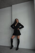 © NASTYA PALEHINA - teen fashion. young girl in black dress-jacket and over the knee boots stands casual with hands on waist and looks apart with smile near white studio wall background. lifestyle concept, free space