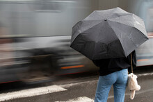 Young Woman In Jeans Under Black Umbrella On A Crossroad On A Rainy Day With Traffic In Motion Blur,  Ear View