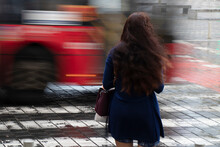 Young Woman With Long Wavy Hair Waiting On A Crossroad On A Rainy Day With Traffic In Motion Blur, Rear View