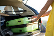© bilanol - Young woman in yellow summer dress taking green suitcase from car trunk. Travel and vacations concept