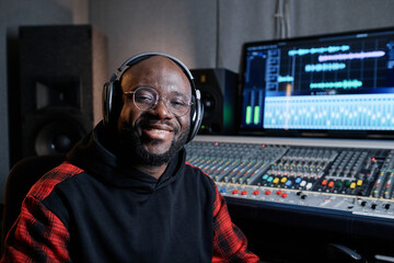 Wall Mural - Portrait of cheerful African American music producer wearing headphones and eyeglasses sitting in recording studio smiling at camera