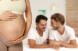 © New Africa - Surrogacy concept. Young pregnant woman and blurred view of happy gay couple indoors