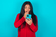 young latin woman wearing red shirt over blue background being deeply surprised, stares at smartphone display, reads shocking news on website, Omg, its horrible!