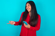 Happy pleased young latin woman wearing red shirt over blue background raises palm and holds cellphone uses high speed internet for text messaging or video calls