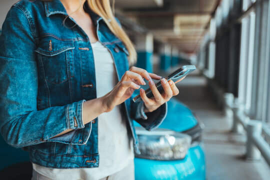 A young Caucasian woman standing nearby car texting message on a smartphone. Attractive brunette with long hair using mobile phone chatting or surfing the internet on the roadside.