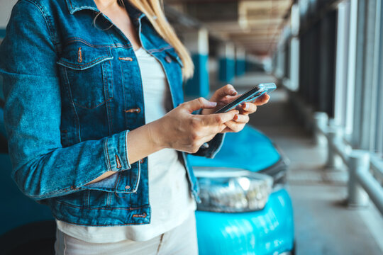 A young Caucasian woman standing nearby car texting message on a smartphone. Attractive brunette with long hair using mobile phone chatting or surfing the internet