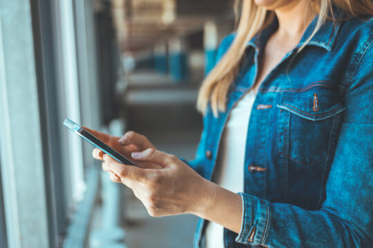 A young Caucasian woman standing nearby car texting message on a smartphone. Attractive brunette with long hair using mobile phone chatting or surfing the internet on the roadside.