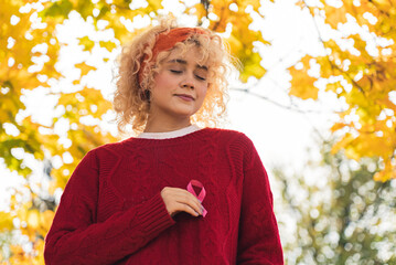 Breast cancer survivor. Outdoor shot of a peaceful caucasian young adult woman holding pink ribbon in front of her chest. Autumn park vibes. High quality photo