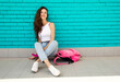teen skater leisure. young fit brunette girl in jeans sits casual with smile on pink skate near backpack on a blue brick wall background and looks happy. lifestyle concept, free space on right side