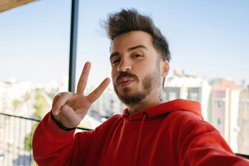 Wall Mural - Handsome young man wearing red hoodie taking a self portrait outdoor at balcony. Happy guy smiling at the camera. Photo of handsome happy young man make selfie by camera. Showing v-sign.