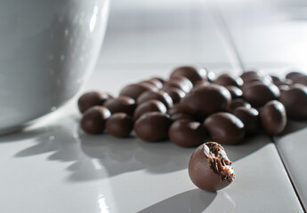 chocolate covered raisins, hazelnuts, coffee beans, almonds pouring out of a white bowl on a white table lit by the sun lighting through the window