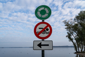 Green sign with a swimming ring, red sign with a crossed out swimmer and white sign with an arrow pointing to the right on a post at the lake beach