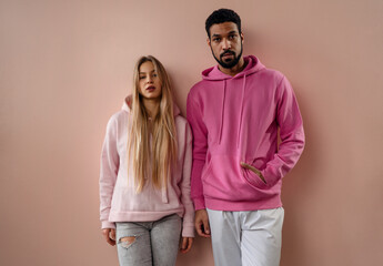 Wall Mural - Fashion studio portrait of young biracial couple in hoodies posing over pink background.